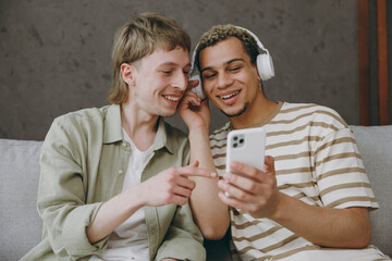 Young couple two gay men wear casual clothes headphones listen music use mobile cell phone sit on...