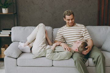 Side view young couple two gay men in casual clothes together lying on partner knees sit on sofa couch at home flat rest spend free spare time in living room. Pride day june month love lgbtq concept