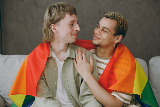 Young smiling happy couple two gay men wear casual clothes wrapped in flag together sit on sofa couch at home flat rest spend free spare time in living room. Pride day june month love lgbtq concept.