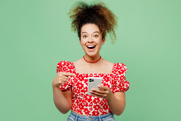 Young surprised shocked happy woman she wear casual clothes red blouse hold in hand use point index finger on mobile cell phone isolated on plain pastel light green color background studio portrait