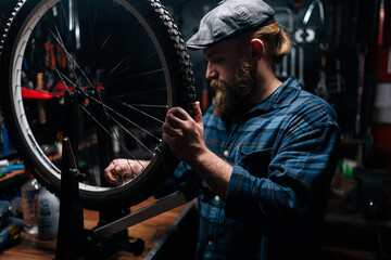 Fototapeta na wymiar Bearded cycling mechanic male in cup checking bicycle wheel spoke with bike spoke wrench working in repair shop with dark interior. Concept of professional repair and maintenance of bike transport.