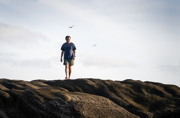 Man walking barefoot on rocks at Muriwai beach with gannets flying overhead. Auckland.