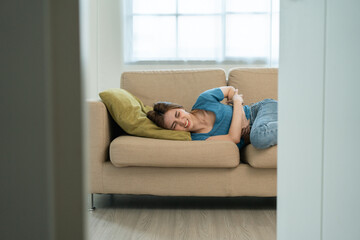 Young asian sick woman hands holding pressing lower abdomen. Medical or gynecological problems. Young woman suffering from abdominal pain on sofa at home healthcare concept. Stomach ache