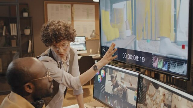 Tilt shot of female Caucasian producer looking and pointing at big and small screens giving male African American colleague recommendations on project