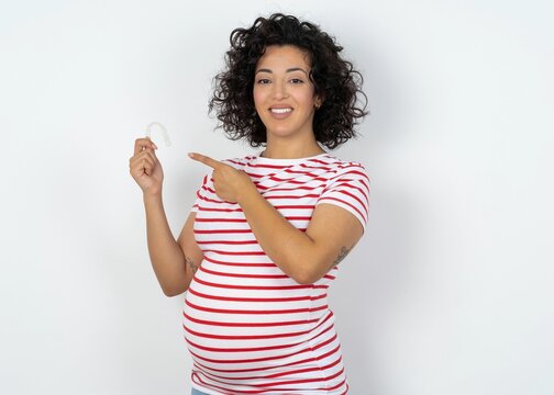 Beautiful pregnant woman wearing striped T-shirt standing over white studio background holding an invisible aligner and pointing at it. Dental healthcare and confidence concept.