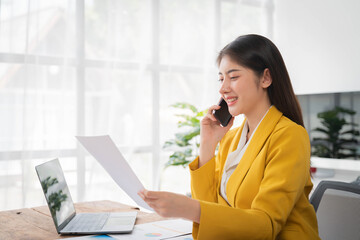 Businesswoman talking on cellphone holding paper sitting at desk and using laptop computer working online in office. Entrepreneurship and business career, distance freelance job concept.