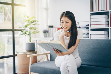 Attractive young Asian woman  sitting on the sofa in the minimal and comfortable living room enjoying reading a book.