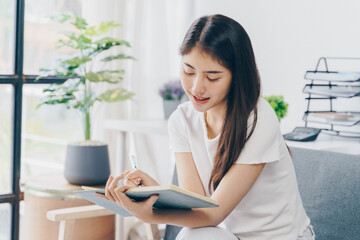 Attractive young Asian woman sitting on the sofa in the minimal and comfortable living room enjoying reading a book.