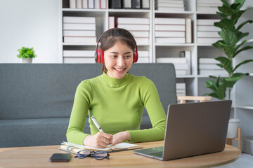 Young asian woman in wireless headphones studying online, using laptop and taking notes while sitting in living room at home.