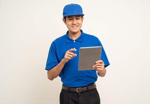 Asian Man In Blue Uniform Standing Holding Digital Tablet Computer On Isolated White Background. Male Service Worker With Cell Phone. Delivery Courier Shipping Service