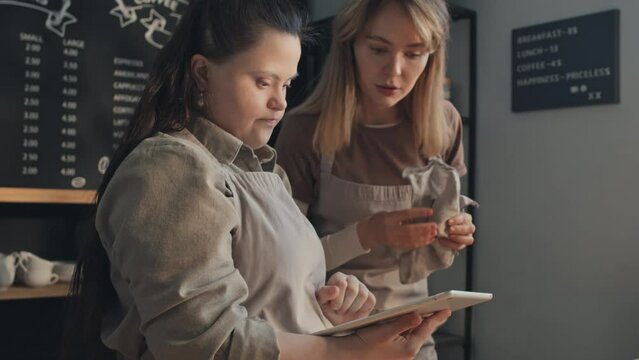 Selective focus shot of young cafe worker checking if her new colleague with Down syndrome know how to use app on digital tablet for work