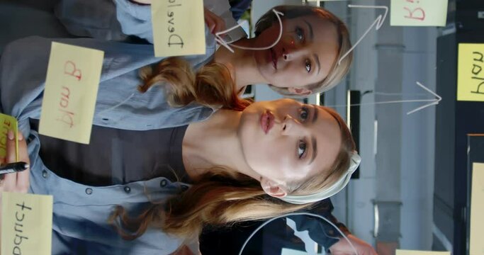 Vertical shoot. Workers gathered in front of transparent board and stick notes. Employees are attracted to work discussion. Young girl in front manages process. Team work at office.