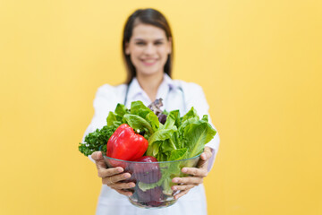 Happy cheerful female doctor or nutritionist showing a vegetables in transparent bowl to camera close up, female doctor holding vegetable basket portrait on yellow.