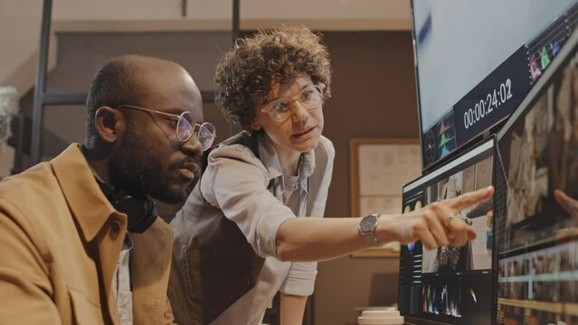 Low angle of female Caucasian producer pointing at screen giving recommendations to male African American editor sitting at desk in office in evening