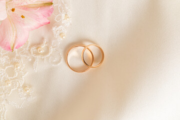 Top view of two gold wedding rings on a satin fabric background with a embroidered with tulle and a pink flower. Wedding background. space for text.