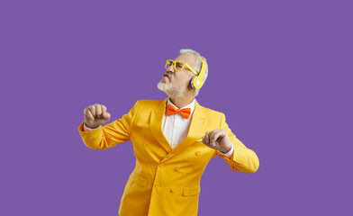 Elderly gray-haired man in glasses for party listens to music in wireless headphones and performs energetic dance making funny face dressed in yellow jacket and bow tie located in purple studio