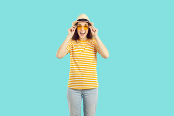 Happy woman in casual clothes enjoying summer holidays. Smiling excited beautiful young girl wearing panama hat, orange sunglasses and striped tee shirt standing isolated on turquoise blue background