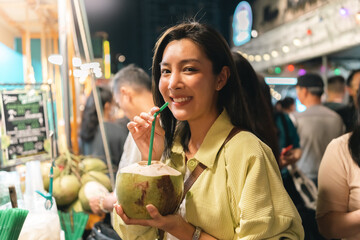 Obraz premium Asian women enjoy drinking coconut water street food at night market. Traveler Asian blogger woman Happy tourists Beautiful female with Traditional thailand bangkok food.