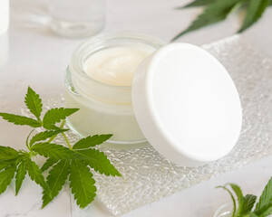 Obraz na płótnie Canvas Opened white cream jar with a lid near cannabis leaves. Cosmetic Mockup