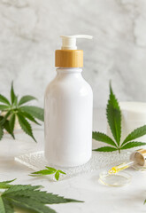 Obraz na płótnie Canvas White One pump bottle near green cannabis leaves on white table. Cosmetic Mockup