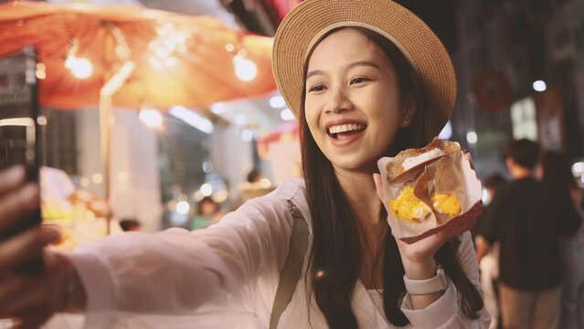 Asian woman tourist backpacker doing selfie and recording a video blog while eating food from street stall in night market with crowd of people at Yaowarat road, Bangkok