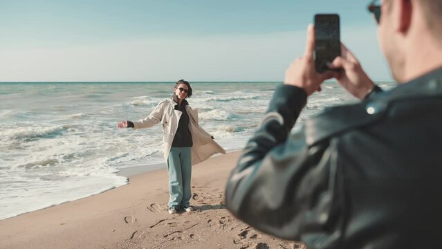 Handsome man taking pictures of girl with his phone camera on beach by sea