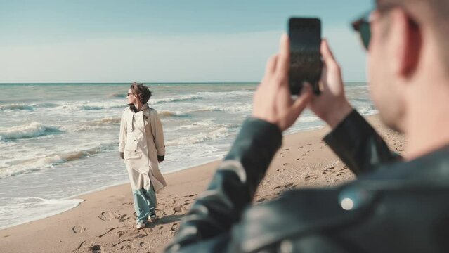 Lovely man taking pictures of girl with his phone camera and hugging on beach by sea