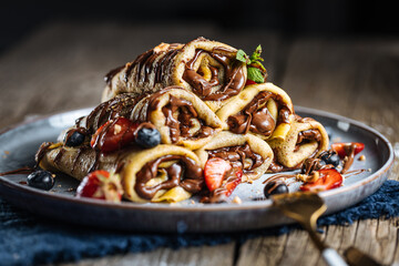 Crepes with chocolate filling arranged in pyramid shape with seasonal fruit like strawberries, cherries and blueberries on a blue plate placed on rustic wooden board