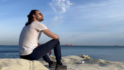 a man in a white t-shirt and glasses sits near the blue sea