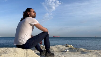 a man in a white t-shirt and glasses sits near the blue sea