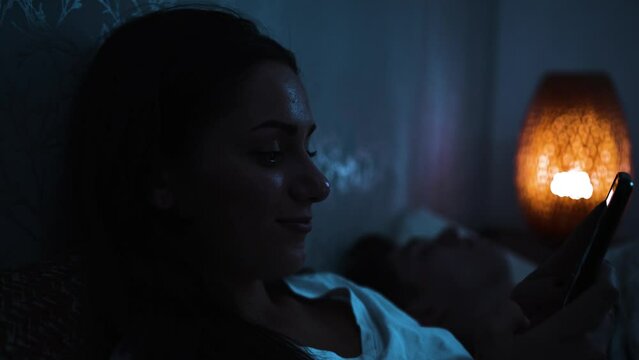Young woman smiles whilst texting on her phone in bed at night