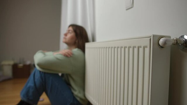 Home radiator with blurred young woman sitting on floor at background rubbing arms talking. Side view unrecognizable Caucasian lady warming up at home indoors