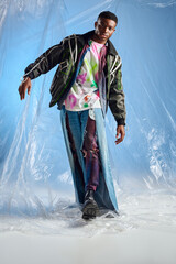 Trendy young afroamerican man in outwear jacket with led stripes and ripped jeans walking and looking at camera on glossy cellophane on blue background, urban outfit, DIY clothing