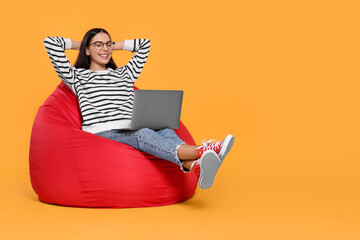 Happy woman with laptop sitting on beanbag chair against orange background