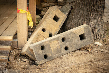 Heavy concrete weight blocks with various holes for temporary construction fence lying near wooden construction and a tree trunk on sand - Powered by Adobe