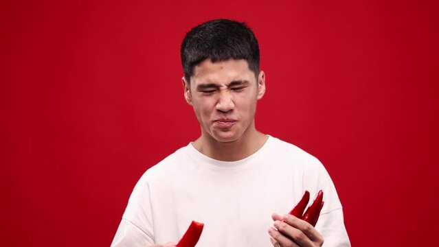 Grimace face. Young asian man eating red hot chilli peppers against red studio background. Crying, spicy hot taste. Concept of food, vegetables, taste, grocery, human emotions. Ad