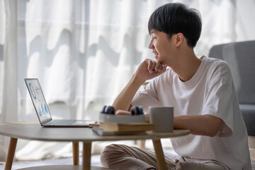 A happy and smart young Asian male college student in casual clothes is doing his homework at a coffee table in his living room.