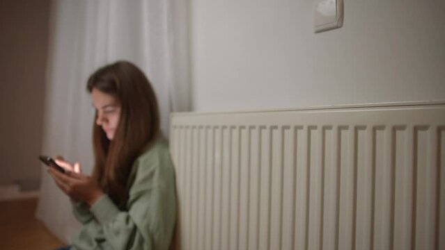 Calefaction radiator in flat with blurred young woman sitting at background on the left scrolling social media app on smartphone. Caucasian lady at heater at home indoors
