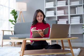 Beautiful young Asian woman sitting using a laptop and take note at home. Young Asian woman student sitting on floor working with laptop computer and taking notes at home.