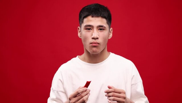 Burning mouth. Young asian man eating red hot chilli peppers against red studio background. Crying, spicy hot taste. Concept of food, vegetables, taste, grocery, human emotions. Ad