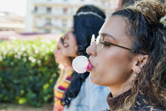 Youthful Energy and Playfulness in a Public Park. Caucasian woman blowing up a chewing gum balloon, surrounded by friends enjoying the outdoors.