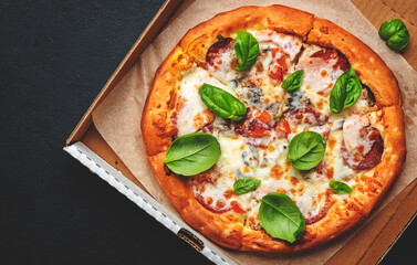 Hot pizza in box with spicy salami sausage, mozzarella cheese, tomato sauce and green basil, just delivered, black table background, top view