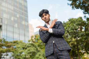 A stylish guy in a blue checkered suit in a serious face with a forbidden gesture while standing outside, in the city. Sky, building and trees are shown in the background.