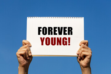 Forever young text on notebook paper held by 2 hands with isolated blue sky background. This message can be used as business concept about forever young.