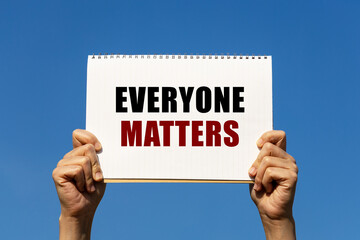 Everyone matters text on notebook paper held by 2 hands with isolated blue sky background. This...