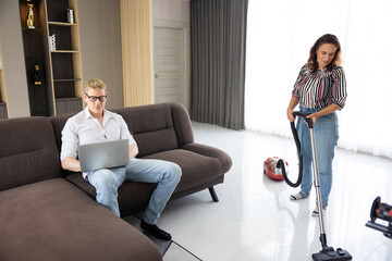 wife doing housekeeping clean house while man working on laptop computer sitting on sofa in living room at home