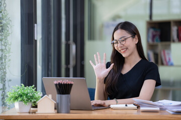 Happy young Asian businesswoman waving hands to greeting partner during making video conference with her team.