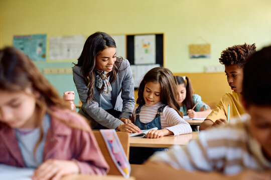Happy Hispanic teacher assists schoolgirl during class at elementary school.