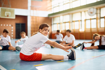Smiling schoolgirl stretching her leg during physical activity class and looking at camera.