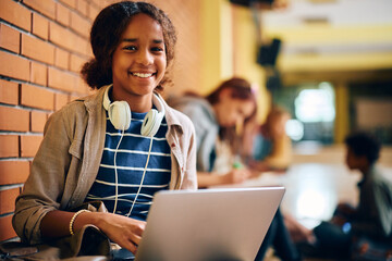 Happy black teenage girl using laptop in high school hallway and looking at camera.